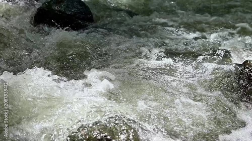 Close-up of powerful rushing river water flowing rapidly over mossy stones. Slow motion shot of crystal clear water splashing in a wild mountain stream.