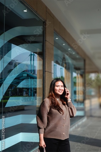 Smiling woman walks outside a modern glass building. She holds her phone, engaged in a pleasant conversation. Her outfit is cozy and stylish, adding warmth to the urban setting.