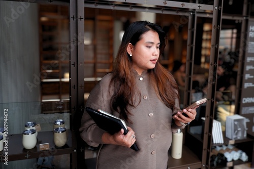 Woman smiles while talking on phone. She holds a tablet and walks through a cafe. Background features shelves and menu boards. Her expression shows friendly, confident engagement.