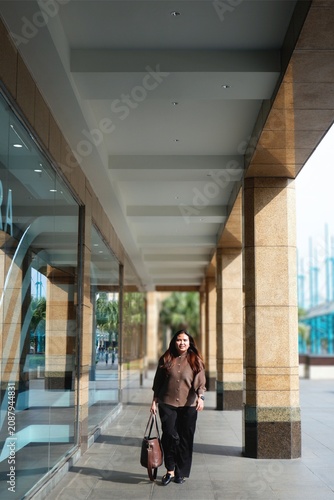 Woman smiles warmly under glass-and-concrete walkway. She holds a leather bag, standing calmly beside pillars. Sunlight spills softly through the open sky above.