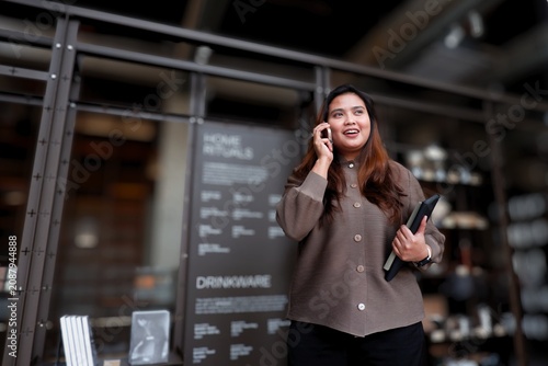 Woman smiles while talking on phone. She holds a tablet and walks through a cafe. Background features shelves and menu boards. Her expression shows friendly, confident engagement.