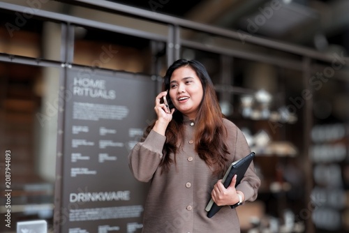 Woman smiles while talking on phone. She holds a tablet and walks through a cafe. Background features shelves and menu boards. Her expression shows friendly, confident engagement.