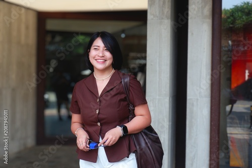 Warm smile lights up the woman's face. She wears glasses and a brown blouse. Her arms cross as she leans against a modern wall. Large bag rests on her shoulder. Her gaze invites connection, warmth.