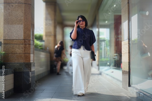 Woman walks through a sunlit urban corridor. She wears a dark top and flowing white trousers. Her hand rises to shield her eyes from bright light. Her expression shows calm focus as she moves forward.