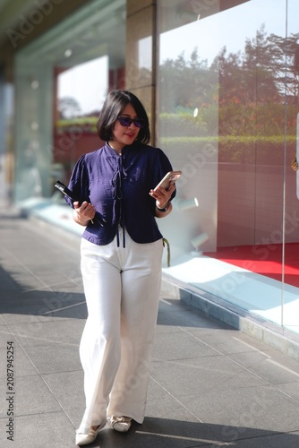 Woman walks through a sunlit urban corridor. She wears a dark top and flowing white trousers. Her hand rises to shield her eyes from bright light. Her expression shows calm focus as she moves forward.