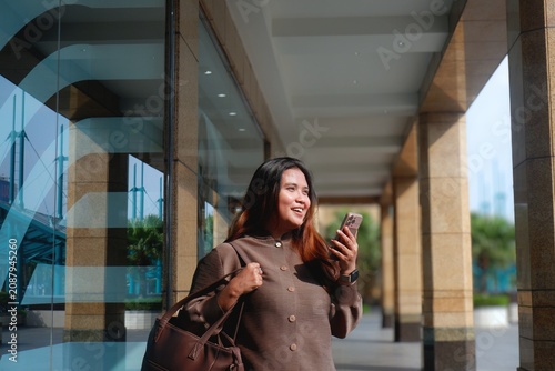 Smiling woman walks outside a modern glass building. She holds her phone, engaged in a pleasant conversation. Her outfit is cozy and stylish, adding warmth to the urban setting.