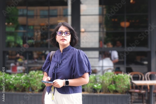 Young woman walks urban street, holding phone. Wearing glasses, blue blouse, and white pants. Casual, confident, focused on her path. Background shows modern cafe with glass windows.