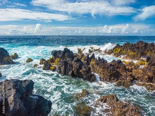 Powerful white ocean waves foaming and crashing into dark volcanic rock reefs near Porto Moniz natural pools, Madeira, Portugal.