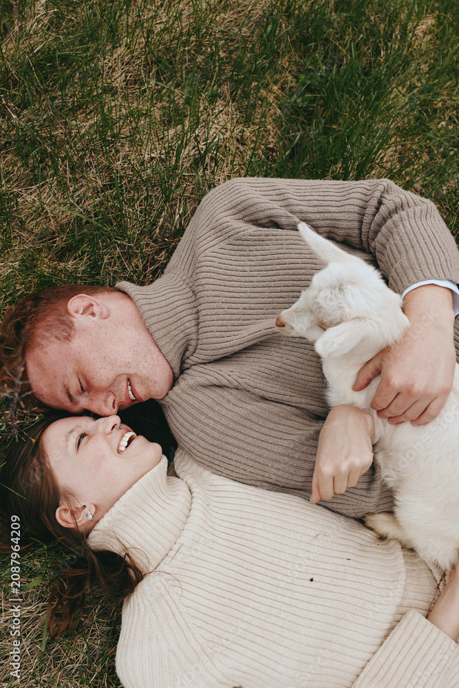 Fototapeta premium A joyful couple lies on grass, cuddling a goat kid, sharing a tender outdoor moment, surrounded by warm sunlight and soft sweaters, capturing simple, everyday happiness and connection.