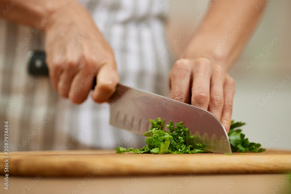 Fototapeta premium Chef's hands holding a knife, finely chopping fresh green parsley herbs on a rustic wooden cutting board in a kitchen
