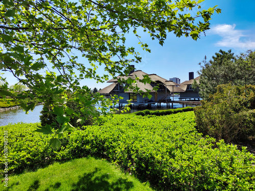 Scenic view of waterfront houses surrounded by green trees, shrubs and manicured lawn. Lakeside landscape with vibrant foliage and blue sky. For travel, architecture, resort outdoor lifestyle.