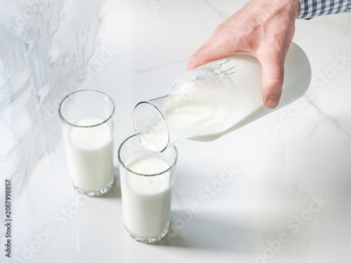 Obraz na plátně Man's hand pours fresh milk from a glass bottle into a clear glass on a white marble table