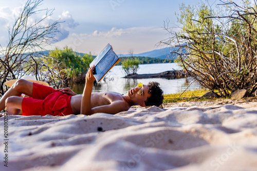 Young man relaxing on beach reading book enjoying the summer vacation by lake