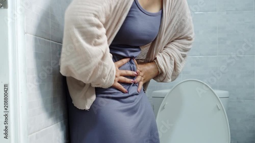 woman suffering from severe stomach ache, food poisoning, or diarrhea, holding her painful belly in front of a toilet bowl in the bathroom. Healthcare and medical concept
