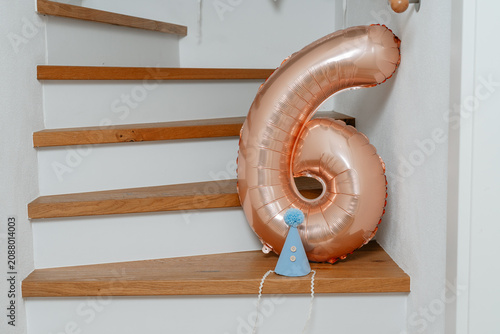 number six balloon resting on wooden stairs with blue party hat above, celebrating birthday party in indoor setting with white walls and brown steps, above view.
