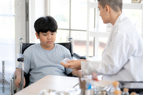 Caucasian woman doctor talking with young asian boy patient sitting in wheelchair for examining at clinic, practitioner explaining medical treatment in hospital, helping little kid with disability.