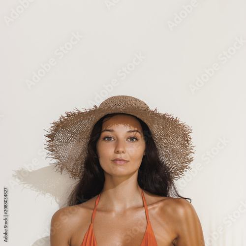Tanned Woman In Straw Hat And Bikini Posing Against Minimalist Backdrop