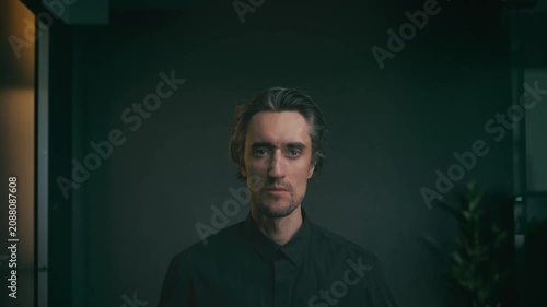 Young man portrait in black button-down shirt, tousled hair, low-key lighting with dramatic shadows, dark indoor background, faint plant silhouette, slight smile