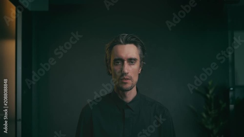 Moody indoor portrait of a man with medium-length hair in dark button-up shirt, standing against dim blurred background with subtle plant, minimalist professional look