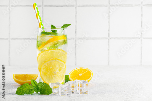 Refreshing homemade lemonade served in clear glass with fresh lemon slices, mint leaves, ice cubes and paper straws on a light kitchen counter against a white tile background with copy space.