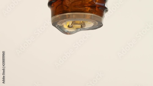 A drop of essential oil slowly moves from a brown bottle, beige background.