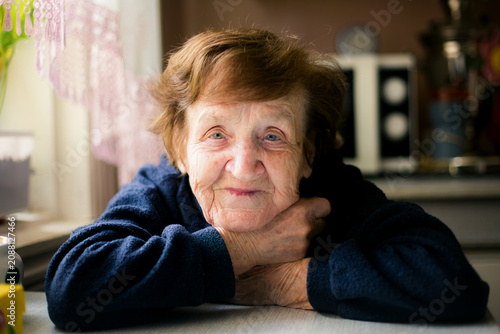 Obraz na plátne Close portrait of an elderly woman resting her folded arms on a kitchen table, illuminated by soft window light in a quiet home interior