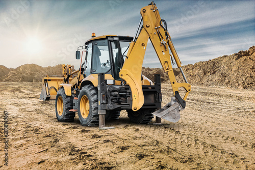 A universal excavator loader is moving dirt at a construction site as the sun shines. Mounds of earth are visible all around the machinery