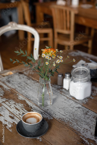 Cup of cappuccino on a rustic wooden table with a small bouquet of wildflowers in a glass vase. Cozy café atmosphere with warm natural light, visible film grain and an analog-inspired vintage aestheti