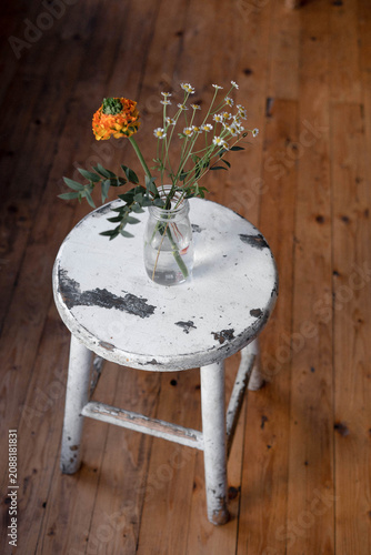 Minimalist still life with a small bouquet of wildflowers in a glass vase placed on a rustic white wooden stool. Warm natural light, visible film grain, analog-inspired aesthetic and cozy home interio