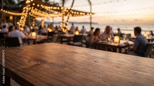 Close-up of an empty dark wood table with a blurred background of a cozy outdoor beach restaurant lit by warm string lights at sunset.