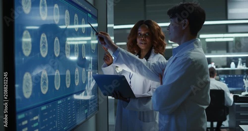 Black Female Neurologist and Asian Male Physician Meeting and Talking, Pointing at MRI Brain Scans on a Large Touch screen Display in a Modern Hospital Laboratory Neuroscience Drug Therapy.