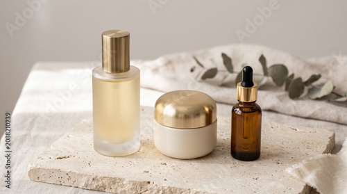 Elegant skincare set with frosted glass bottle, cream jar, and amber dropper on a stone slab with eucalyptus, featuring gold accents and soft light.