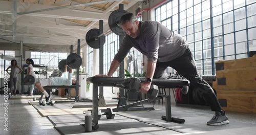 Middle-aged man in long sleeve top preparing on flat bench, picking up hex dumbbell for strength