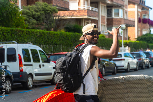 Cuadro en lienzo Young black man carrying surfboard and backpack signaling shaka on city street