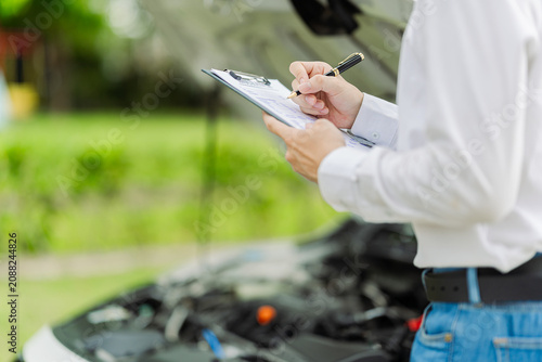  Car insurance agent inspecting engine under open hood and taking notes on clipboard. Professional technician or adjuster checking vehicle engine condition after accident.