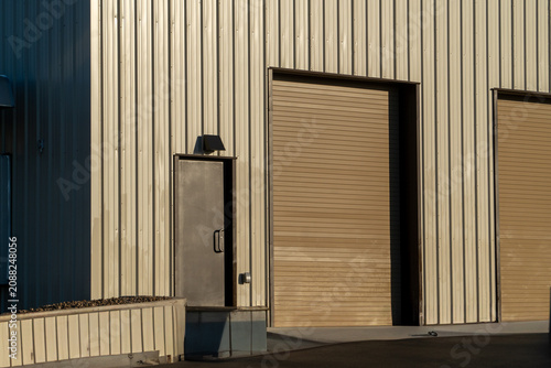 Фотография Industrial building exterior with beige corrugated metal siding, a dark loading dock access door, and a large brown roll-up garage door, bathed in warm sunlight creating dramatic shadows
