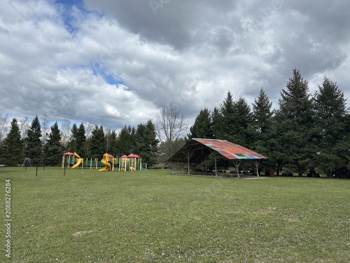 Playground area with colorful play structures and a large shelter surrounded by green grass and tall trees under a cloudy sky in a public park setting
