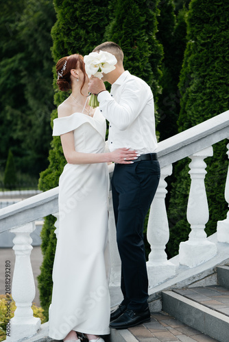 Obraz na plátně A romantic shot of a newlywed couple kissing while playfully hiding their faces behind a beautiful bouquet of white calla lilies on a balcony stairs