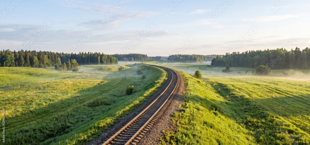 Naklejka premium A solitary railway track curves through a misty green meadow and dense forest under a clear blue sky at dawn