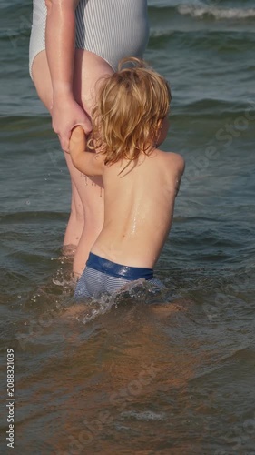 Young child seen from behind holding adult hand while stepping into shallow seawater, with splashing droplets, ripples, foam, and bright coastal light creating family beach scene