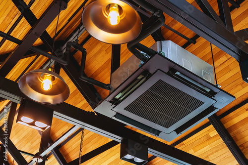 Modern cassette type air conditioner installed on loft ceiling with steel beams and wooden roof. Ceiling-mounted cassette air conditioning unit integrated into modern interior design. HVAC systems.