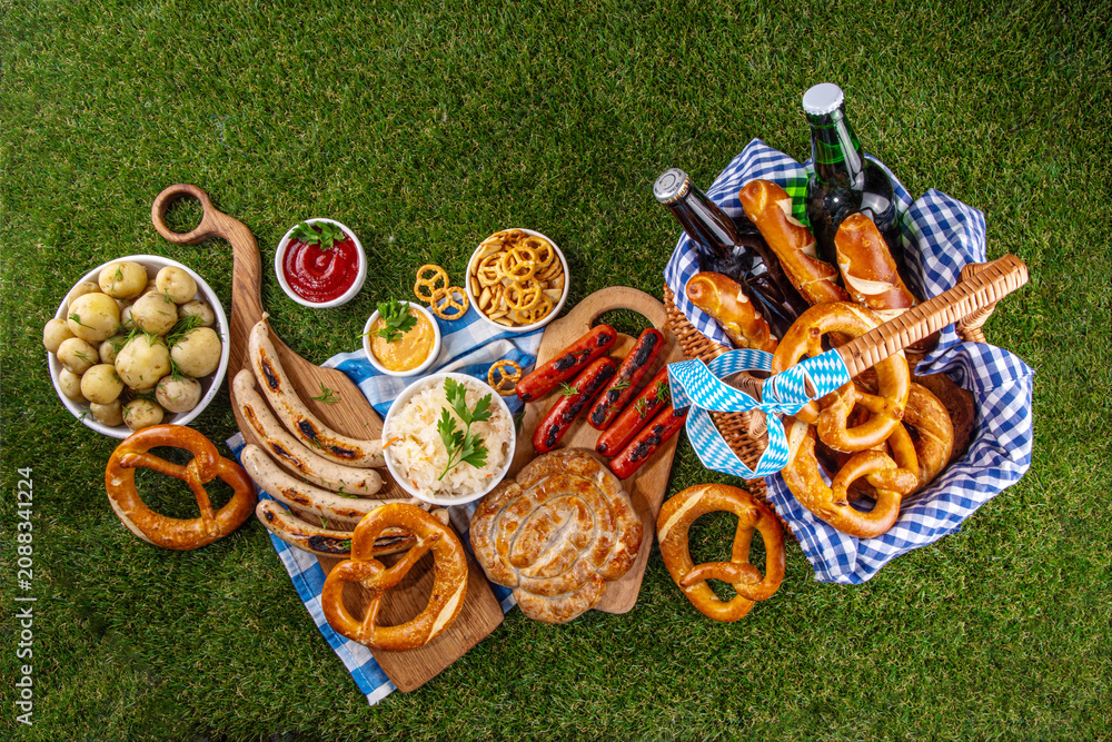 Fototapeta premium Oktoberfest picnic and barbecue food, bavarian party menu, bavarian sausages, pretzels, potato, sauerkraut, beer bottles, with picnic basket, checkered napkin tablecloth, Oktoberfest festive ribbon