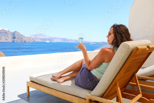 Woman relaxing on a sunlit Santorini terrace, seated on a cushioned lounge chair overlooking the caldera, enjoying wine and the serene blue Aegean during a peaceful Greek island escape.