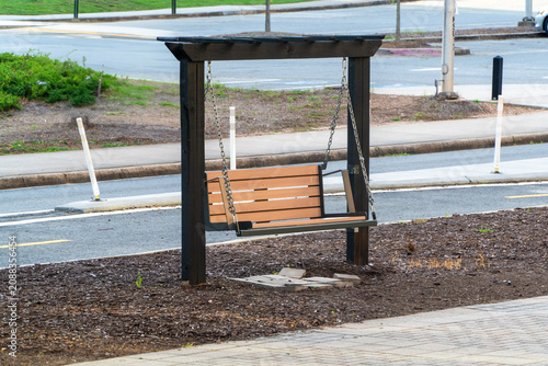 Fotografia Modern wooden swing bench with a dark frame and light slats, providing a peaceful resting spot in an urban park or garden, surrounded by mulch, paved path, and a quiet street