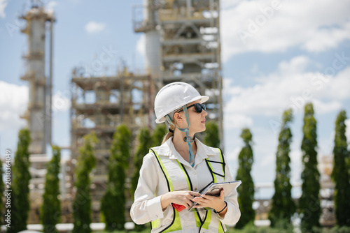 Female engineer in hard hat observing construction site with industrial structures in background under clear blue sky