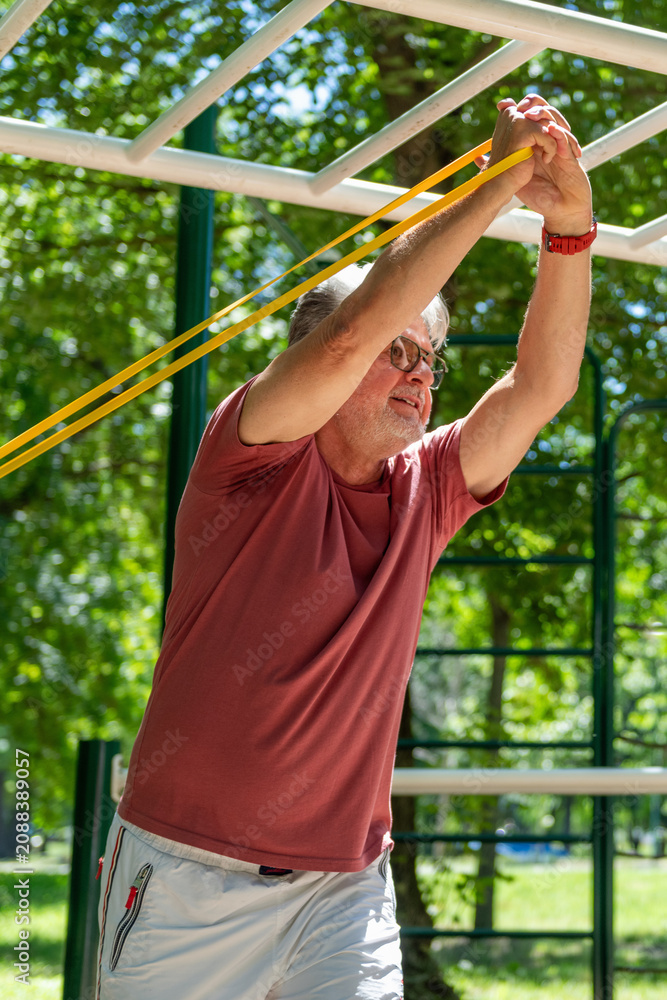 Obraz premium Senior man performing an overhead resistance band exercise in an outdoor fitness area to enhance shoulder mobility, flexibility, posture, and functional movement.