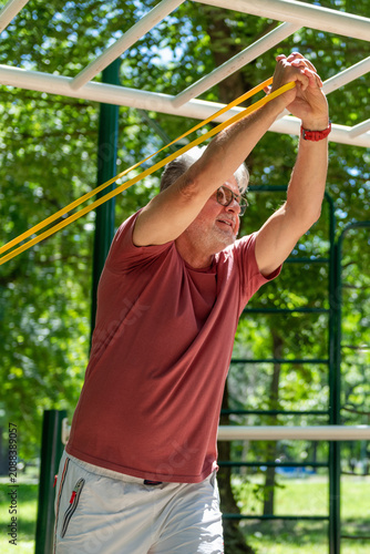Senior man performing an overhead resistance band exercise in an outdoor fitness area to enhance shoulder mobility, flexibility, posture, and functional movement.