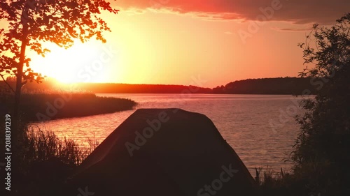 Camping tent pitch on riverbank during vivid and intense crimson sunset in Kyiv region, Ukraine, featuring rich red and orange tones in sky reflecting on water.