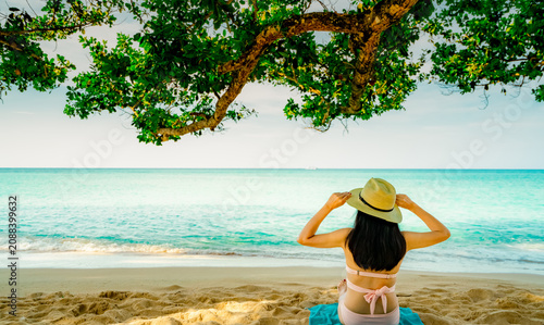 Obraz na plátně Back view of a woman wearing a straw hat relaxing beneath a tropical seaside tree