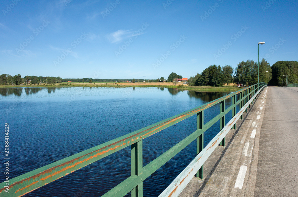 Fototapeta premium Bridge over the Dalälven river south of Hedesunda in Sweden, with calm water, green fields, and a clear blue sky